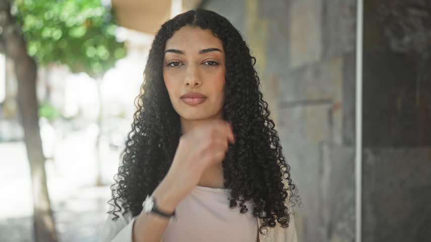 Young woman touching her lip with fingers, wearing wristwatch and casual shirt, standing by building on street; confidence poise.