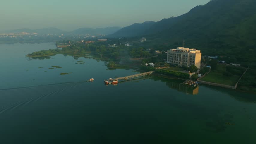 Aerial view of a hotel on the shore of Lake Pichola with a boat on the water and mountains in the background in Udaipur, Rajasthan, India.