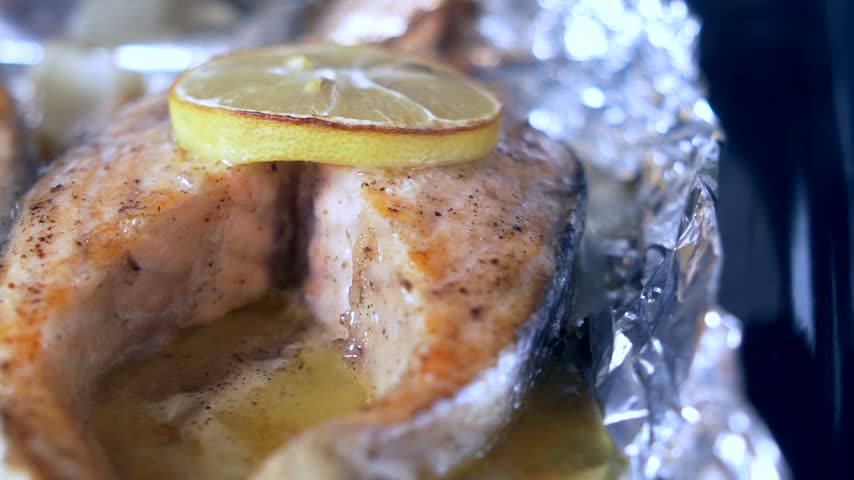 Macro view of oven roasted salmon fish fillets topped with citrus lemon slices and herb butter sauce resting on silver aluminum foil for healthy dinner preparation concept.