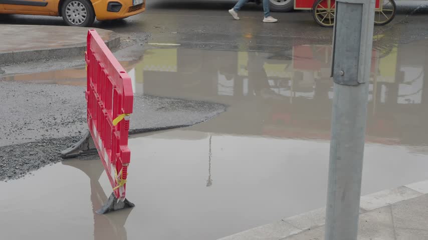 Portable red plastic construction barrier positioned near large water puddle on asphalt surface. Rain day in urban environment with moving cars and blurry street background scene.