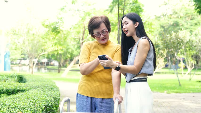 Mature woman using a walker with support from a younger woman in a park, representing elderly care, mobility assistance, rehabilitation, and family support.