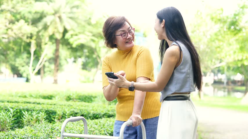 Mature woman using a walker with support from a younger woman in a park, representing elderly care, mobility assistance, rehabilitation, and family support.