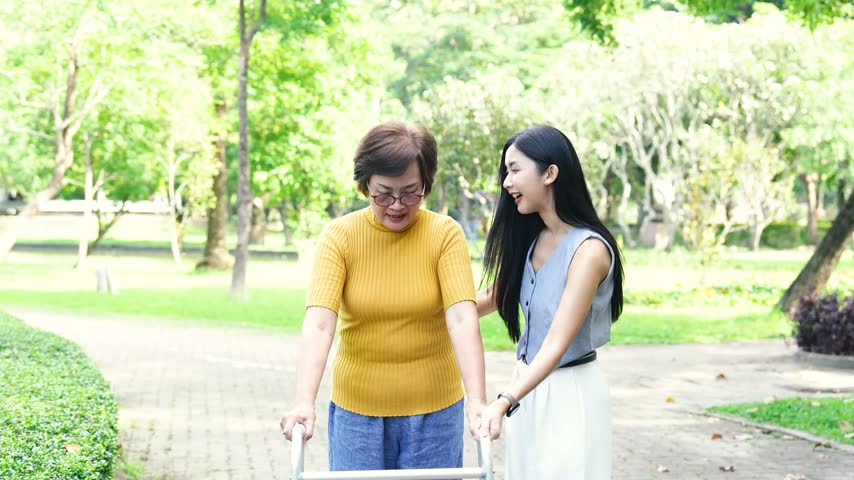 Mature woman using a walker with support from a younger woman in a park, representing elderly care, mobility assistance, rehabilitation, and family support.