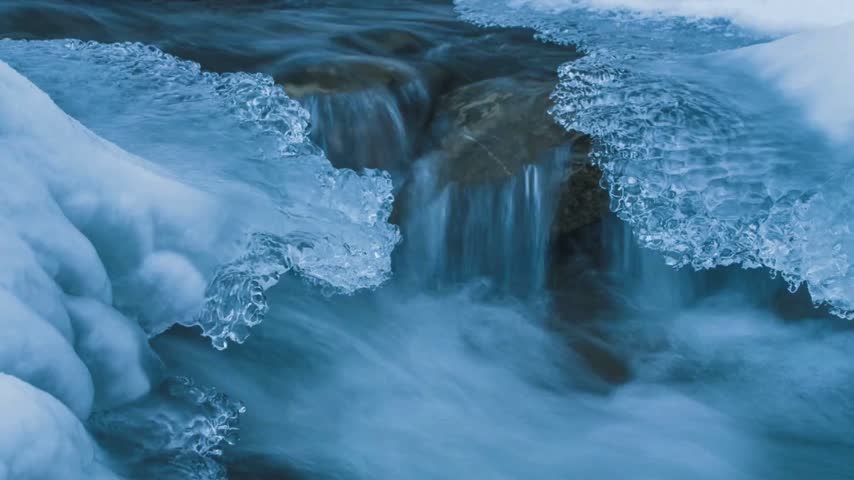 Crystal clear water flows over dark river rocks framed by jagged ice shelves and textured frozen snow in deep winter landscape with cold blue tones and natural light reflecting on moving stream