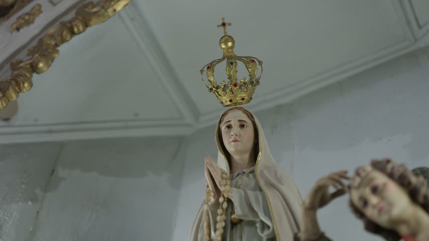 A statue of Our Lady of Fatima wearing a gold jeweled crown and holding a rosary, seen from below in a white chapel.