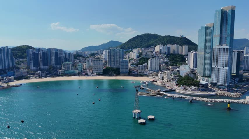 Busan, South Korea - 24 June 2026: Aerial view of Haeundae Beach with modern skyscrapers, the cable car tower in the water, and lush green hills under a clear blue sky.