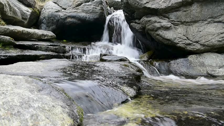 Flowing water tumbles over smooth stones in a rocky terrain amidst lush greenery, showcasing the beauty of nature in a secluded area.