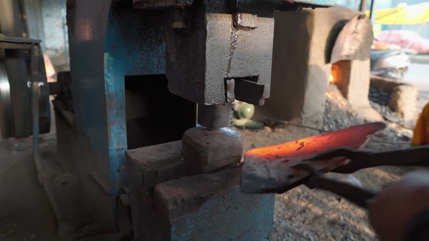 Close-up view of a blacksmith working with heated metal in a metalworking workshop. The worker uses long tongs to position glowing hot metal on an anvil while a mechanical power hammer repeatedly strikes the workpiece. The scene shows hands-on forging, traditional craftsmanship, heavy metalworking equipment, and the intense process of shaping heated metal.