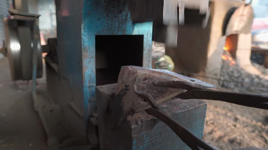 Close-up view of a blacksmith working at an anvil, using metal tongs to hold a workpiece beneath a heavy forging hammer. A traditional forge with visible fire and glowing embers appears in the background. The rustic workshop setting highlights manual metalworking, traditional craftsmanship, forging techniques, and the transformation of metal using heat, tools, and mechanical force.