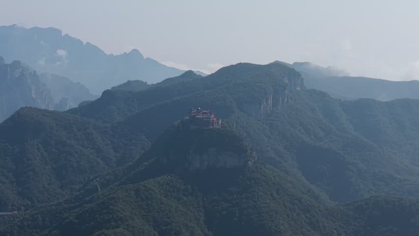 Aerial view of a traditional red temple on a solitary peak in Yuntai Mountain, Jiaozuo. Surrounded by lush green forests, deep valleys, and misty Taihang ridges under a hazy sky. Serene travel landscape footage.