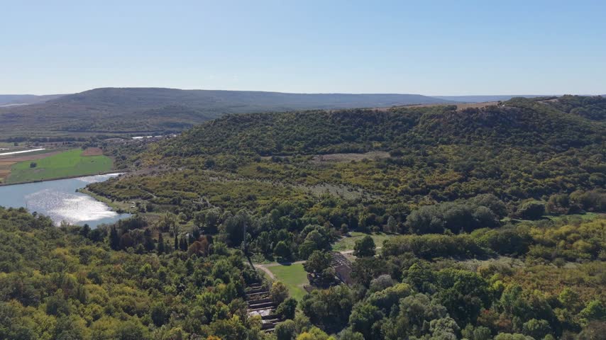 Scenic drone view of lake shoreline, green fields, wooded hills, and distant mountains.