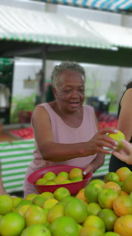 Older Black woman selecting ripe green and yellow oranges at an open-air produce stand, enjoying a familiar neighborhood grocery routine