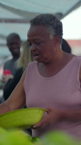 Senior African American shopper holding a bowl at an open-air fruit stand, thoughtfully inspecting fresh oranges during her regular neighborhood grocery run