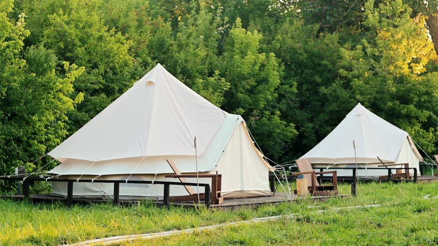 Glamping tents set on wooden platforms with chairs and tables near green forest under bright sunlight.