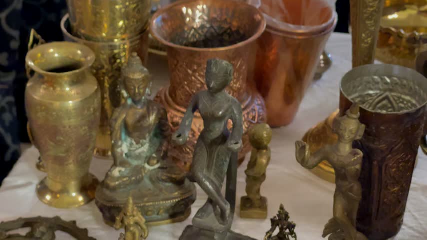 A close-up shot of vintage handcrafted copper and brass statues, featuring a Buddha figurine alongside carved metal idols and engraved copper pots in an antique store display.