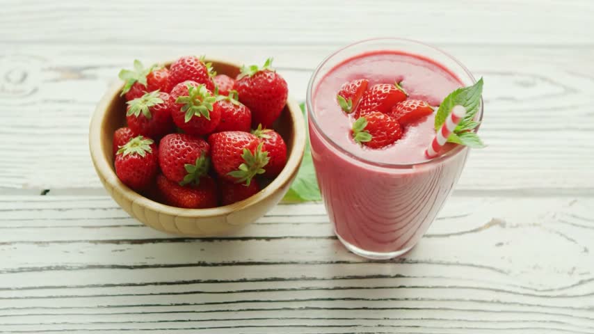Fresh strawberry smoothie drink in glass with bowl of ripe berries on table.