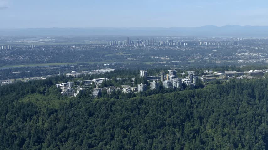 Simon Fraser University Atop Forested Burnaby Mountain With Metro Vancouver Downtown Skyline In The Background In Canada. - aerial shot