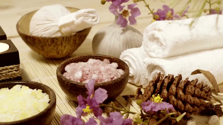 Spa setup with towels, herbal compress, salt scrubs, and candles on a wooden table with fresh purple tropical flowers for tranquil wellness therapy.