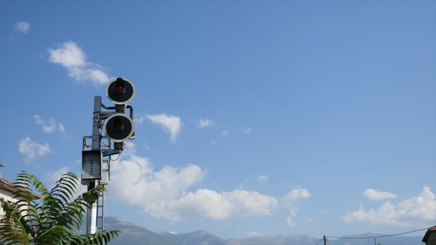 Red railway signal beside the tracks in Sora, Frosinone, Lazio, Italy, with tree leaves moving in the wind and mountains under a blue sky in the background.