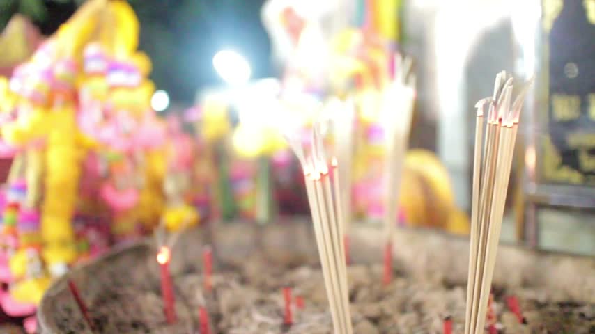 Incense sticks burn in sand pot at Buddhist shrine during night time ceremony with blurred vibrant flower garlands creating spiritual atmosphere in temple sanctuary for traditional religious worship