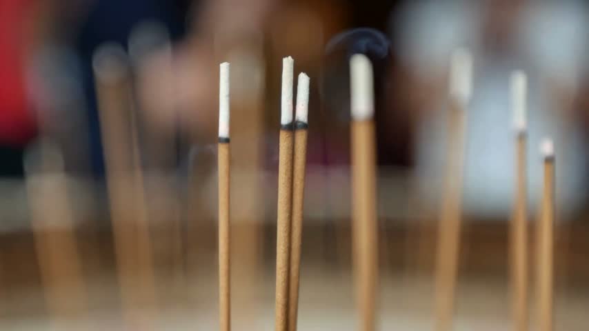 Burning incense sticks inside sand pot at temple with people praying in blurred background creating peaceful spiritual atmosphere for meditation and traditional religious ritual in daily life practice