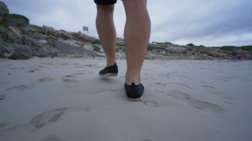 Close up perspective of man walking along sandy shore towards rocky cliffs under cloudy sky creating natural travel atmosphere with outdoors leisure activity at seaside beach during daytime hours