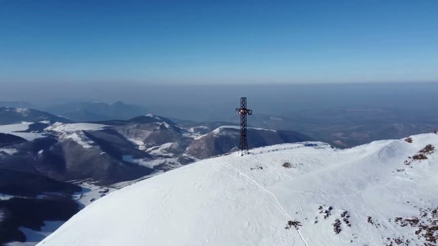 A metal cross sits atop a snowy mountain peak under a deep blue sky near a communication tower, capturing a scenic, high-altitude winter landscape.