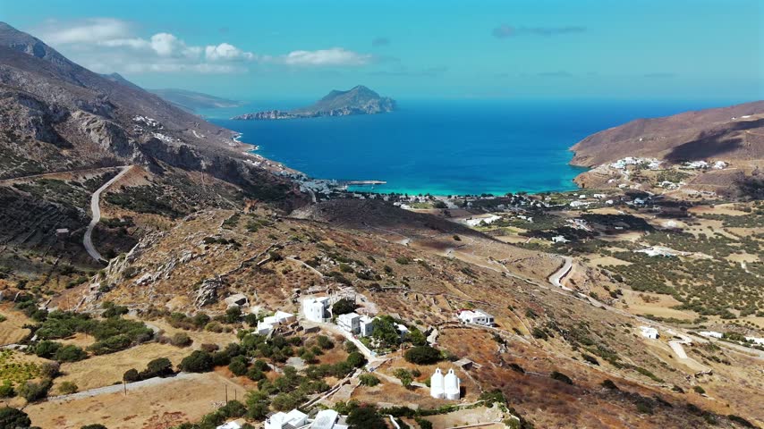Aerial drone shot flying over an arid terraced hillside toward the deep Aegiali bay with turquoise shallows and the mountainous islet of Nikouria on the north coast of Amorgos, Cyclades, Greece