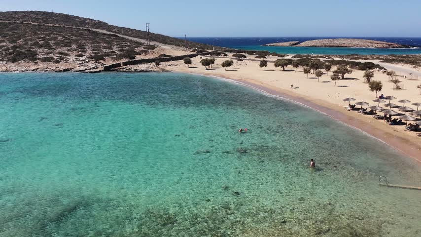 Aerial drone shot of the sandy beach of Kalotaritissa with tamarisk trees and thatched umbrellas beside clear turquoise shallows and a rocky islet across the bay on Amorgos, Cyclades, Greece