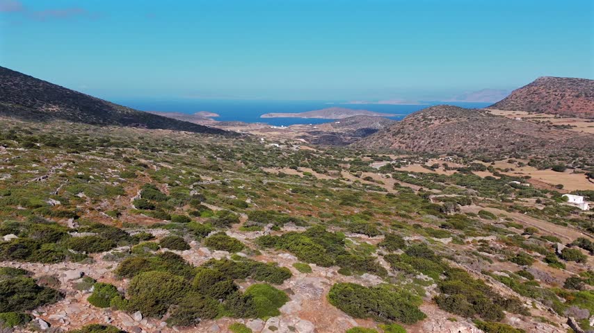 Aerial drone shot over an arid scrubby valley and dry-stone terraces descending toward a distant bay and the blue Aegean Sea on Amorgos, Cyclades, Greece, on a sunny day