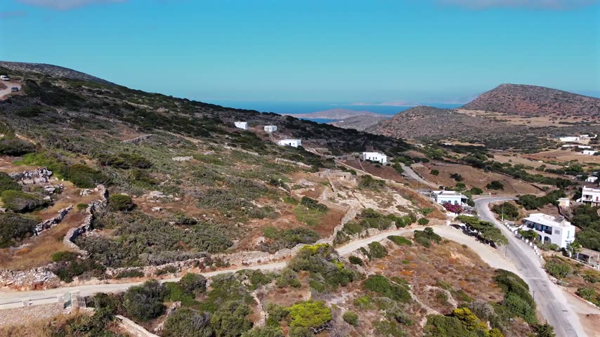 Aerial drone shot of an arid scrubby hillside with scattered white houses and dirt tracks above a distant bay and the blue sea on Amorgos, Cyclades, Greece, on a sunny day