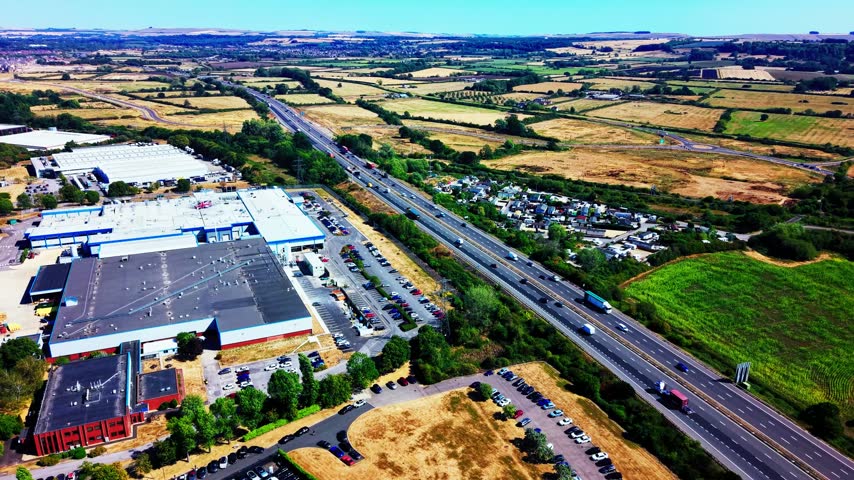Aerial view of the M4 motorway running beside industrial buildings, business premises and surrounding farmland in England with moving traffic and logistics infrastructure
