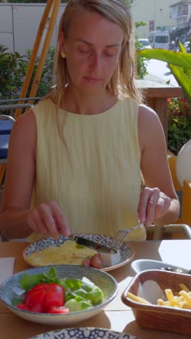Woman having a leisurely breakfast at an outdoor cafe in Fethiye, Turkey, cutting an omelet served with cheese while fresh vegetables and French fries are arranged on the table.