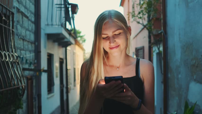 A young blonde woman wearing a black tank top reads her smartphone with a smile while standing in a narrow historic European street during daytime.