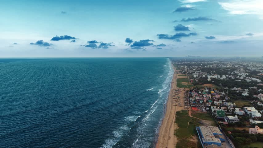 Aerial view of the coastline with sandy beach, blue sea, and urban buildings in Chennai, Tamil Nadu, India.