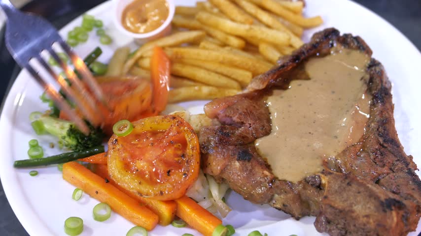 Appetizing t-bone steak topped with savory gravy served with crispy french fries and roasted carrots tomatoes and broccoli on white ceramic plate ready for consumption high angle shot.