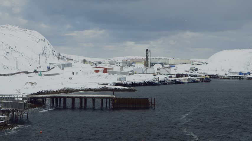 Wide establishing shot of a snowy industrial bay with buildings and docks on the cold coast of the Northern Sea in winter.