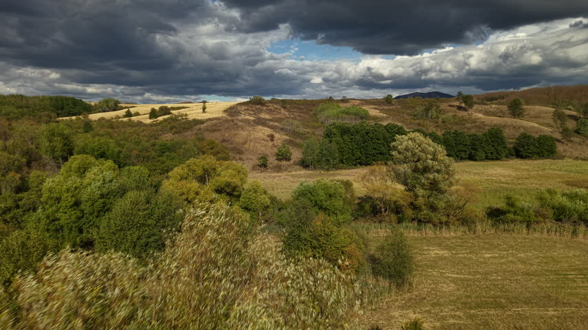 Dramatic storm clouds hang over rolling golden hills and dense green forests in this expansive, moody rural landscape photograph.