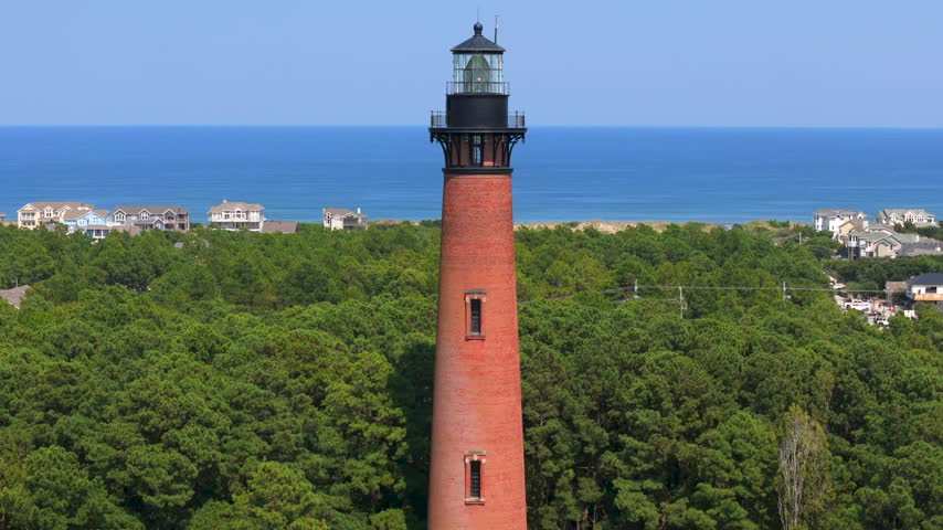 Red Brick Currituck Beach Light Station Stands Above Forested Coastline Overlooking Blue Atlantic Ocean Waters In Corolla, North Carolina, USA. - aerial pullback shot