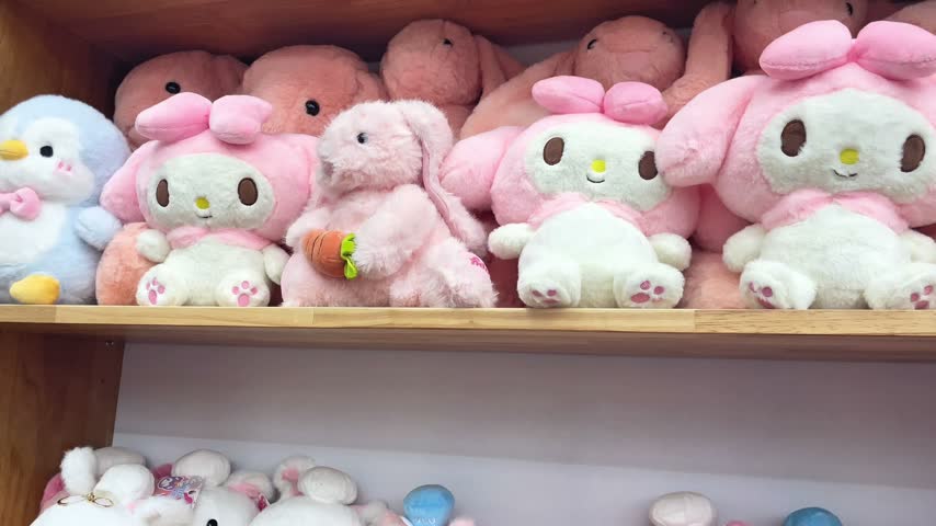 Cute cartoon plush toys and stuffed animals arranged on wooden shelves. A large collection of soft pink piglets with blue accessories and white plush rabbits sitting in rows inside a gift shop. A close-up view of adorable novelty plushies and collectible characters displayed for children or retail customers.