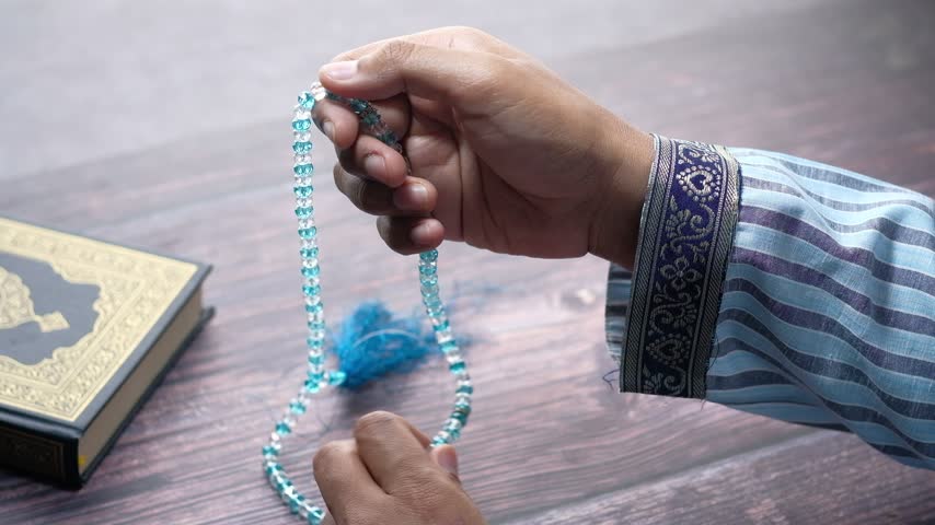 Close up shot of religious man reciting dhikr with blue crystal tasbih beads during islamic prayer session beside book of quran on dark wooden table with soft lighting cinematic footage.