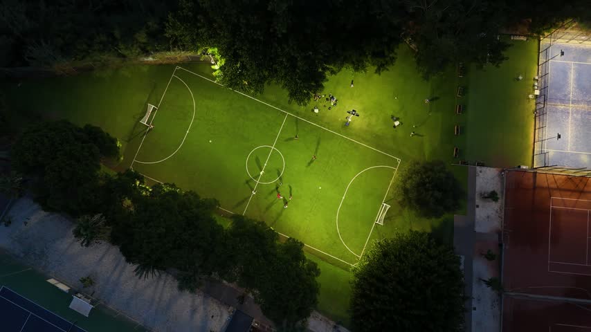 Aerial top-down drone footage of a sports club in Mijas, Costa del Sol, Malaga province, Andalucia, Spain, captured at night. The video shows an illuminated football field with people playing, surrounded by tennis and padel courts, trees, and bright floodlights lighting this recreational sports facility in southern Spain.