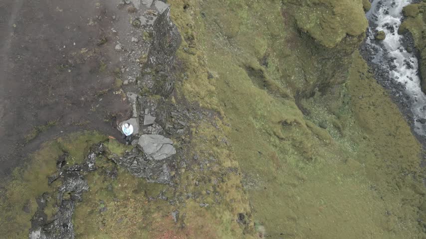 Aerial view of a person standing on a rocky cliff edge overlooking a flowing river in the rugged landscape of ICELAND.