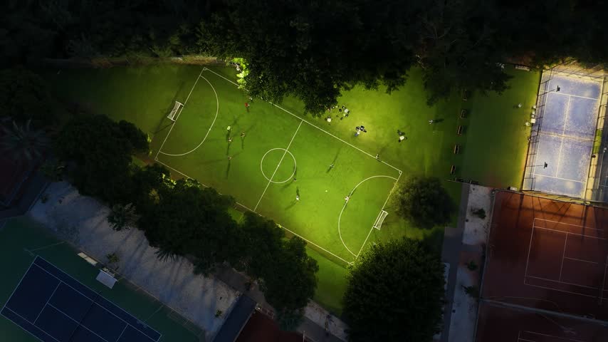 Aerial drone footage with a slow rotating camera over a sports complex in Mijas, Costa del Sol, Malaga province, Andalucia, Spain, captured at night. The orbiting shot reveals an illuminated football field with people playing, surrounded by tennis courts and trees, lit by bright floodlights in this southern Spain community.