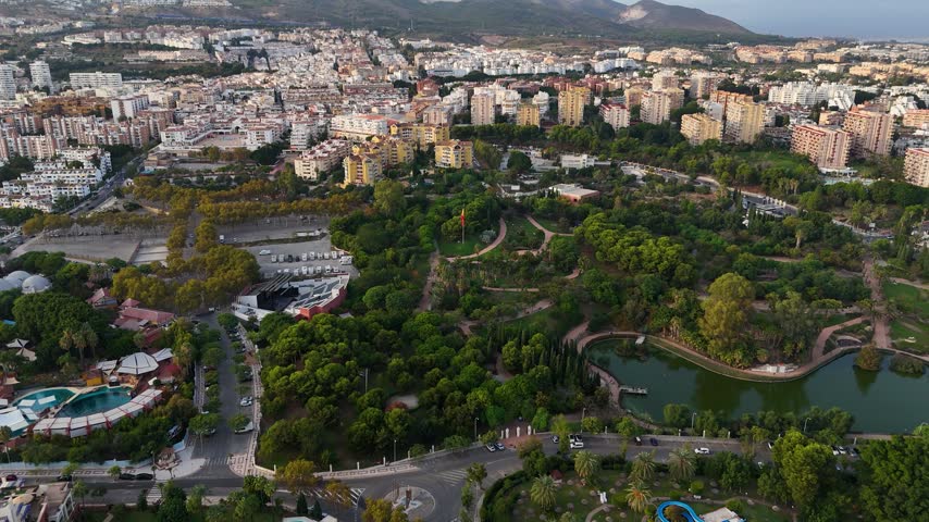 Aerial drone footage of Parque de la Paloma, a public park in Benalmadena, Costa del Sol, Malaga province, Andalucia, Spain. The wide angle view shows the park lake, walking paths, palm trees, and an open air amphitheater, surrounded by apartment buildings with the Mediterranean sea and distant mountains visible in the background.