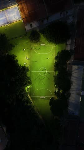 Vertical aerial top-down drone footage of a football field in Mijas, Costa del Sol, Malaga province, Andalucia, Spain, captured at night. The video offers a closer view of the illuminated field with people playing a match, surrounded by tennis and padel courts and tall trees, lit by bright floodlights in this community.