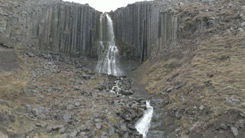 Aerial view of Litlanesfoss waterfall cascading down a dramatic cliff face framed by geometric basalt columns in ICELAND.