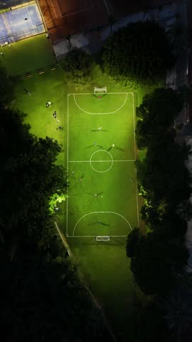 Vertical aerial top-down drone footage of a sports club in Mijas, Costa del Sol, Malaga province, Andalucia, Spain, captured at night. The video shows an illuminated football field with people playing a match, surrounded by tennis and padel courts and tall trees, lit by bright floodlights in this residential community.