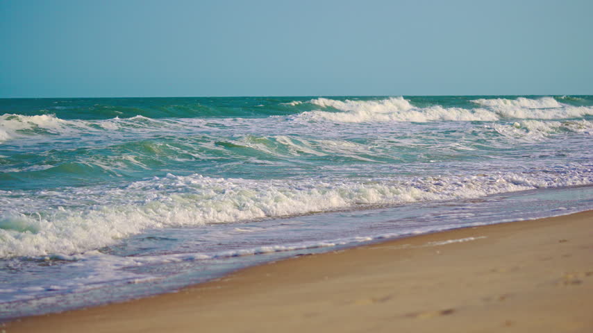 Seaside background with foamy turquoise ocean. Sea beach with sand along tropical coast. Sunny sea beach beside idyllic shore. Ocean waves beneath dreamy blue summer sky.