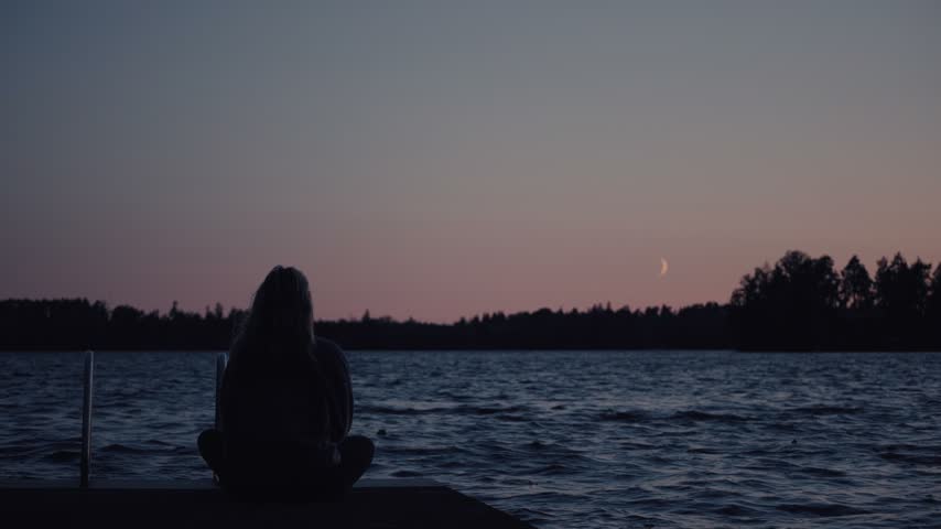 A girl sits quietly on a lakeside pier, watching the sunset and crescent moon over calm water. Serene evening light, tranquil mood, silhouetted figure, natural landscape, scenic view.